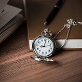 Reloj de bolsillo plateado con cadena apoyado sobre una mesa de madera, junto a un bolígrafo y un libro